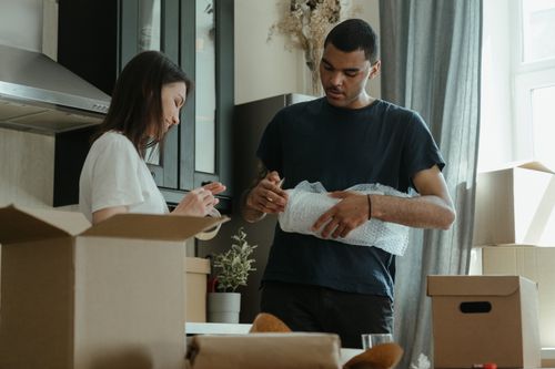 Deux personnes préparent des cartons et emballent des objets dans une pièce lumineuse lors d’un déménagement, avec des boîtes en carton autour.