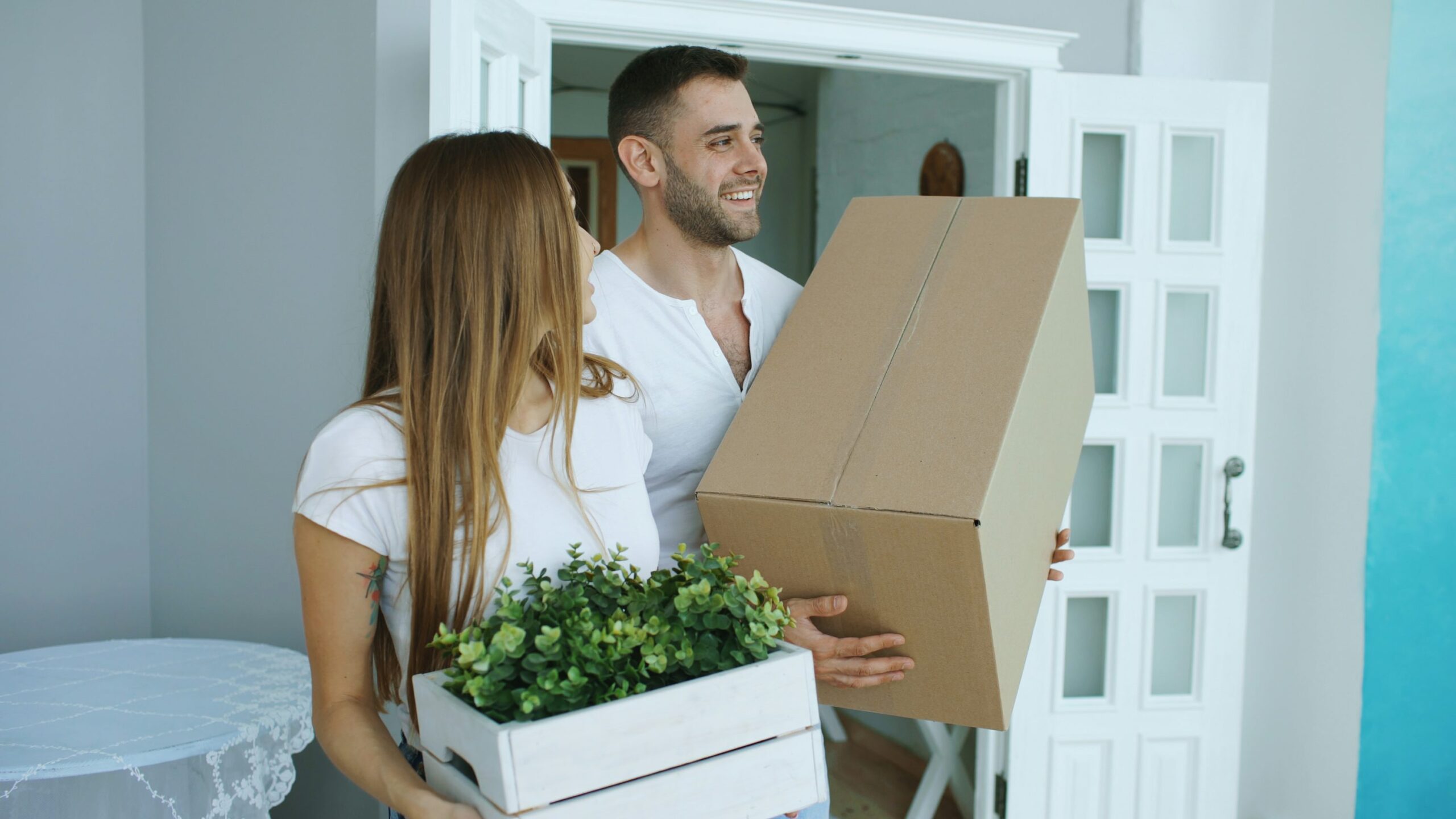 Deux personnes entrant dans une maison avec des cartons et des plantes en main, symbole de déménagement.