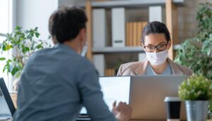 Deux professionnels en réunion de déménagement, l’un face à l’autre avec ordinateur, portables, et masques, dans un bureau. Alt: check-list déménagement professionnel.