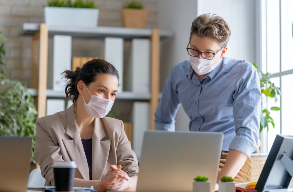 Deux professionnels en salle de travail, masqués, discutent devant un ordinateur portable.