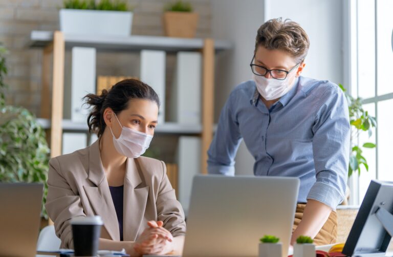 Deux professionnels en salle de travail, masqués, discutent devant un ordinateur portable.